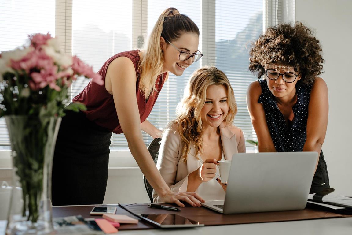 Group of young businesswomen looking at laptop and smiling in office. Three business colleagues working together in modern workspace.